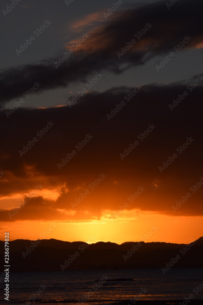 A bright orange sunset nestled between a dark sky and the black hillside in Gisborne, New Zealand.