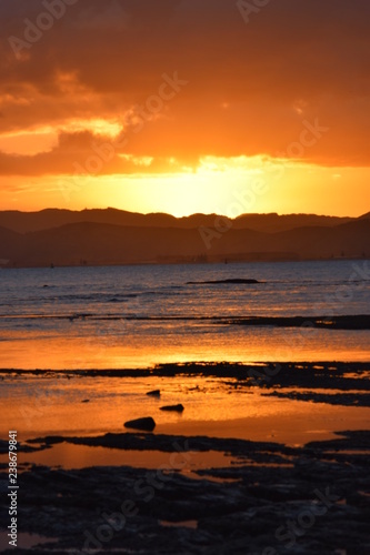 A bright sun setting above the dark hills reflected in the beach shallows at Gisborne  New Zealand.