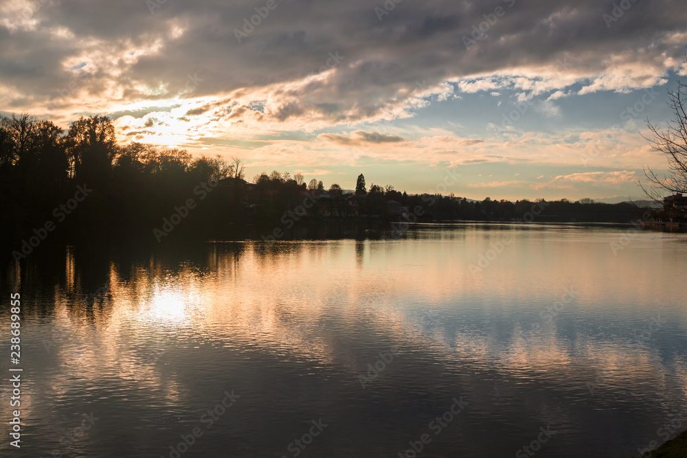 Panoramic view of the Ticino river with clouds and trees that are reflected in its clear water, on a winter day at sunset.