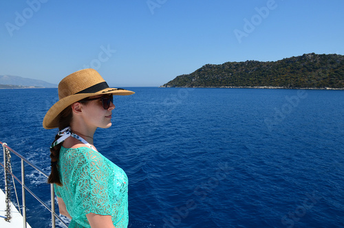 A young brunette girl in a straw hat, sunglasses and a turquoise beach tunic looks away against the background of the sea. © Irina Kulikova