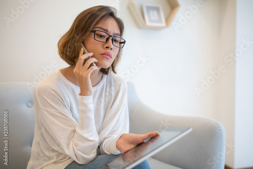 Focused woman holding tablet and talking on phone on sofa. Pretty young Asian lady sitting and using digital devices at home. Technology and communication concept.