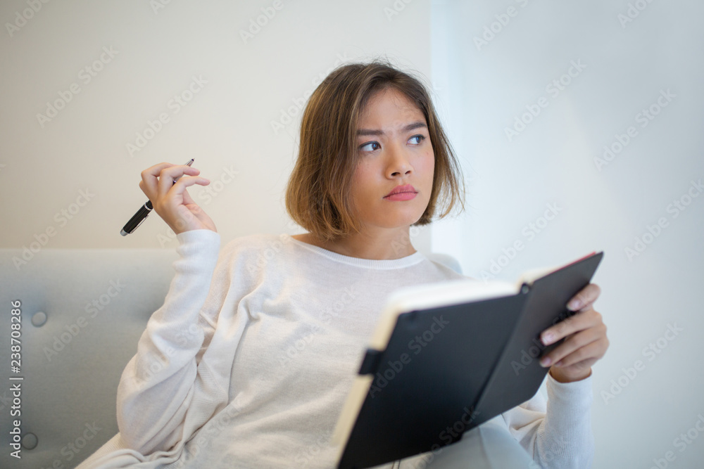 Pensive pretty Asian woman studying at home. Young lady making notes in notebook and sitting on sofa at home. Education or planning concept. Front view. Pensive pretty Asian woman studying at home. Young lady making notes in notebook and sitting on sofa at home. Education or planning concept. Front view.