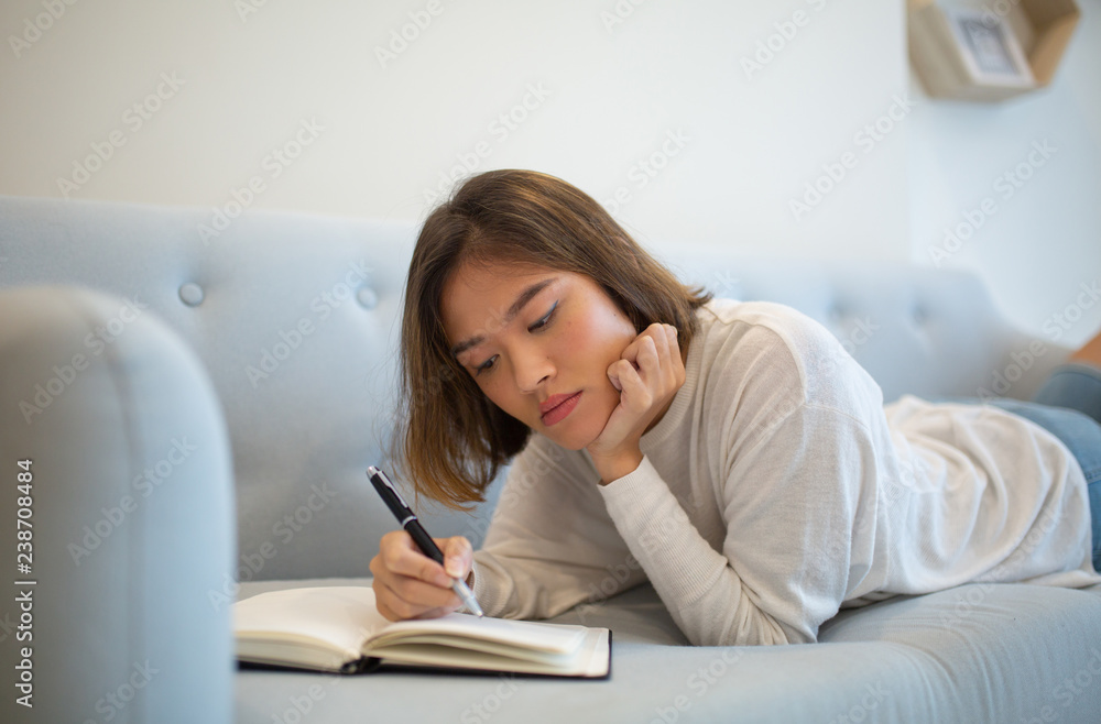 Serious woman making notes in notebook and lying on sofa at home. Pretty young Asian lady studying at home. Education concept. Serious woman making notes in notebook and lying on sofa at home. Pretty young Asian lady studying at home. Education concept.