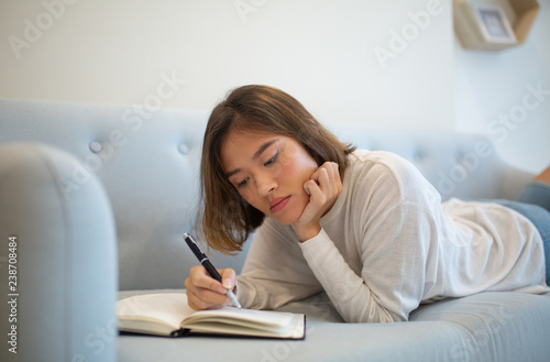 Serious woman making notes in notebook and lying on sofa at home. Pretty young Asian lady studying at home. Education concept.