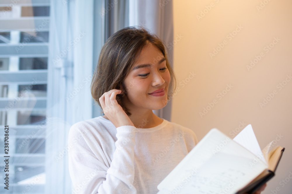 Smiling Asian woman reading book at window at home. Pretty young lady holding book and standing at home with window in background. Education concept. Smiling Asian woman reading book at window at home. Pretty young lady holding book and standing at home with window in background. Education concept.