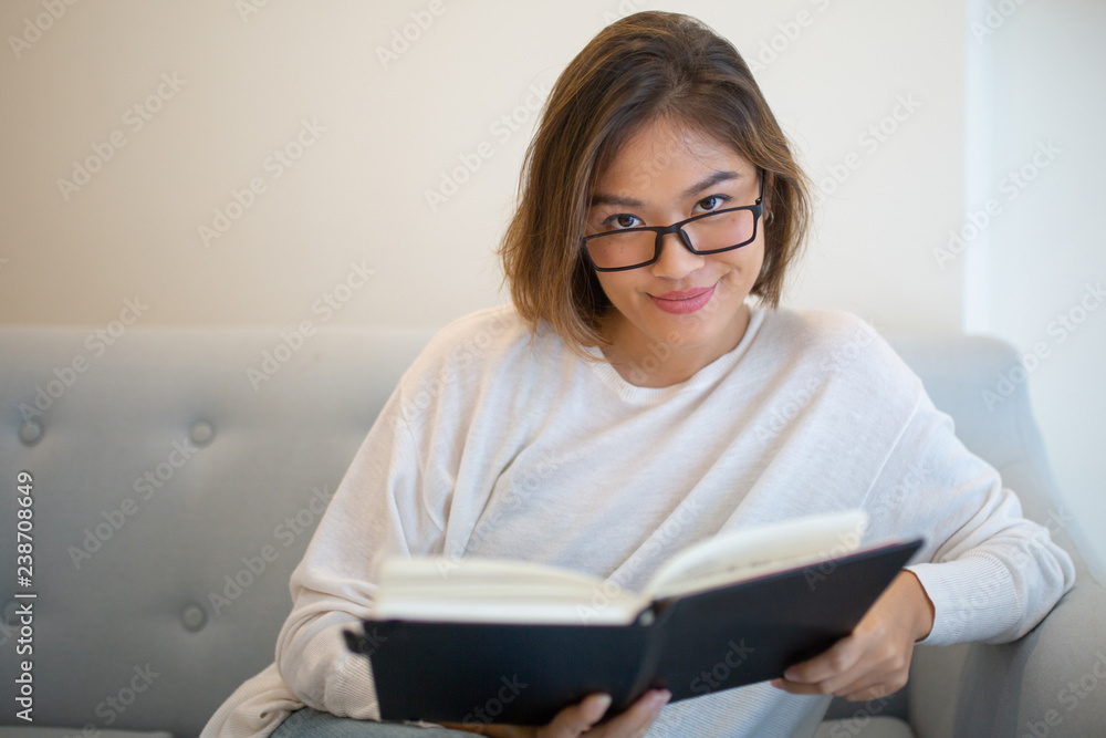 Smiling Asian woman reading book on sofa at home. Pretty young lady looking at camera, sitting and holding book. Education or literature concept. Front view. Smiling Asian woman reading book on sofa at home. Pretty young lady looking at camera, sitting and holding book. Education or literature concept. Front view.
