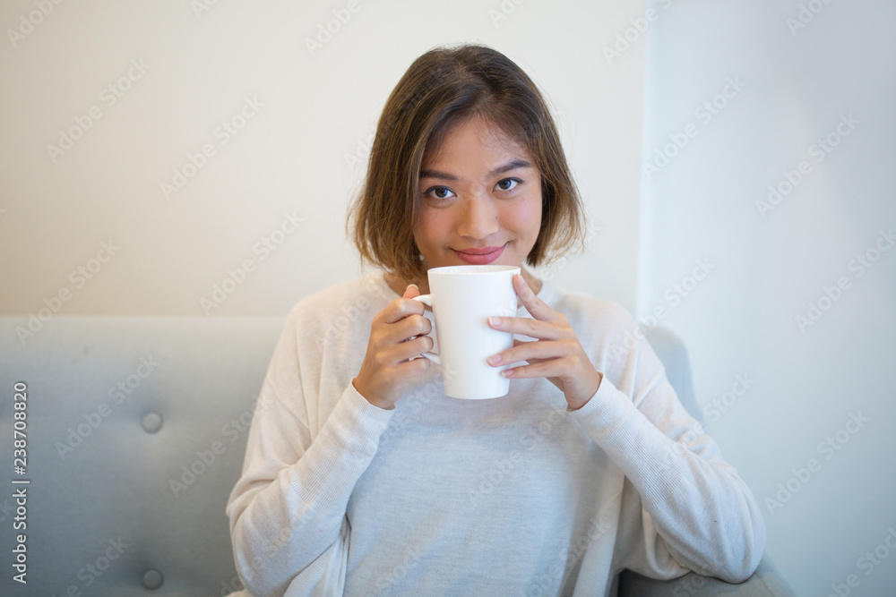 Smiling pretty woman drinking tea and sitting on couch at home. Young Asian lady looking at camera and relaxing. Leisure and tea concept. Front view. Smiling pretty woman drinking tea and sitting on couch at home. Young Asian lady looking at camera and relaxing. Leisure and tea concept. Front view.