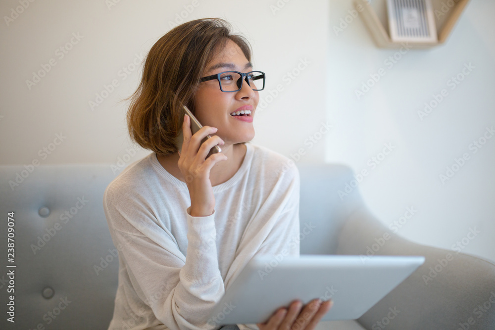 Smiling woman holding tablet and talking on phone on sofa. Pretty young Asian lady sitting and using digital devices at home. Technology and communication concept. Smiling woman holding tablet and talking on phone on sofa. Pretty young Asian lady sitting and using digital devices at home. Technology and communication concept.
