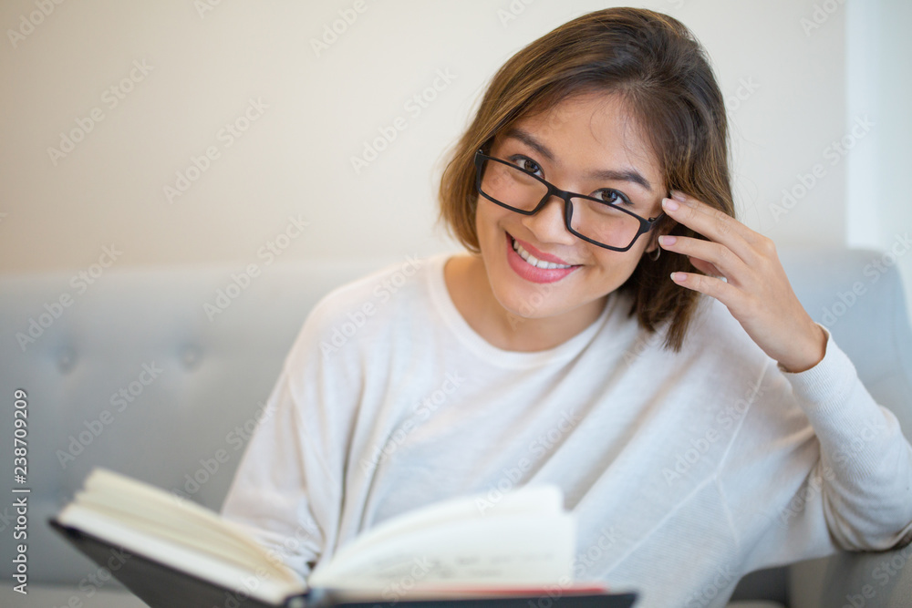 Smiling young woman reading book on couch at home. Pretty Asian lady looking at camera, sitting and holding book. Education or literature concept. Front view. Smiling young woman reading book on couch at home. Pretty Asian lady looking at camera, sitting and holding book. Education or literature concept. Front view.