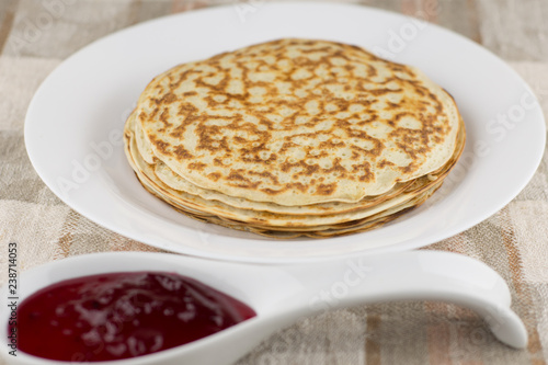 the celebration of carnival: fresh cooked real Russian pancakes made with yogurt, on a white plate and currant jam blurry found. Linen tablecloth, front view, close