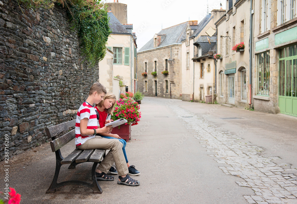 Obraz premium Teenagers travel concept. Children with map sitting on the bench in the street of an old town, historical centre of the town, Rochefort-en-Terre, France