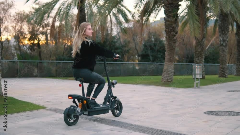 Pretty blonde woman driving electric bike in the park with palm trees. Modern city transportation Pretty blonde woman driving electric bike in the park with palm trees. Modern city transportation