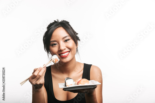 Cheerful young asian woman eating sushi with chopsticks