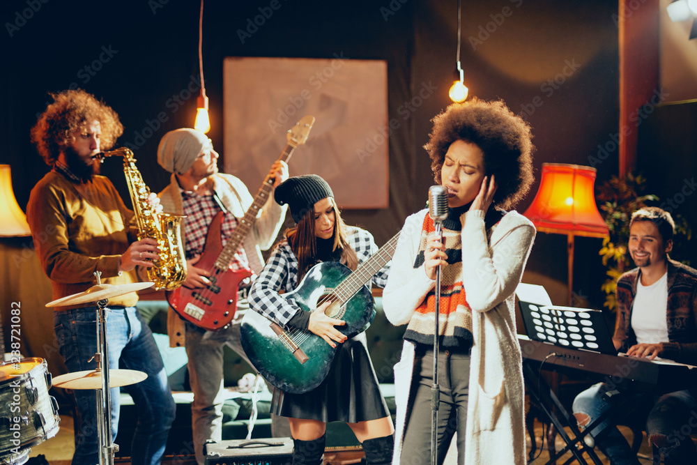Fototapeta premium Mixed race woman singing. In background band playing instruments. Home studio interior.