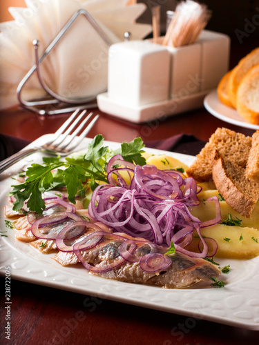 Appetizer of salted herring, red onion, potatoes and rye bread in white plate on dark brown wooden table in restaurant. Vertical