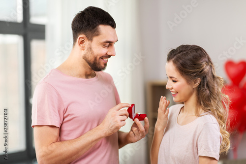 love, couple, proposal and people concept - happy man giving diamond engagement ring in little red box to woman at home