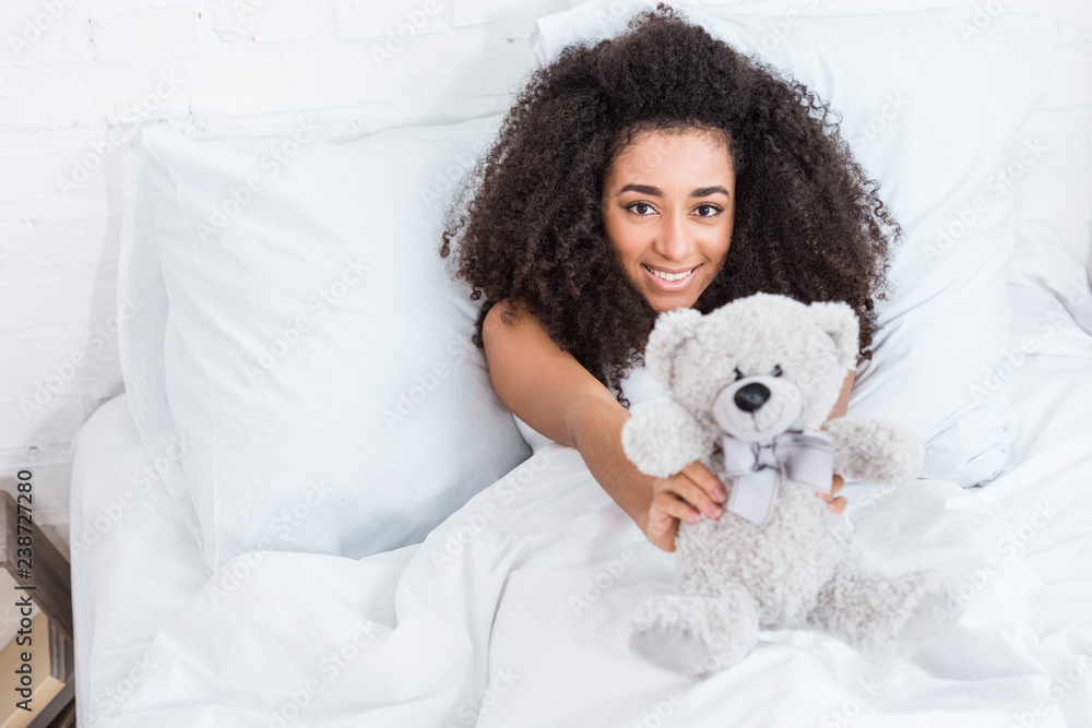 high angle view of of young african american woman laying in bed and showing teddy bear at home