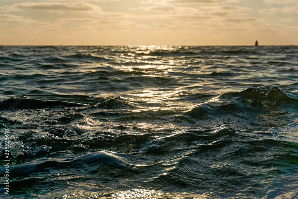 Obraz premium Low view image of waves on the sea at an orange colored sunset and sailing ship on the horizon.