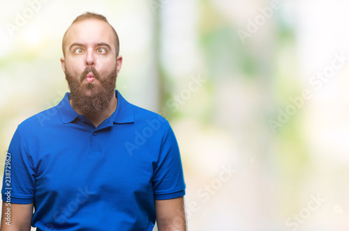 Young caucasian hipster man wearing blue shirt over isolated background making fish face with lips, crazy and comical gesture. Funny expression.