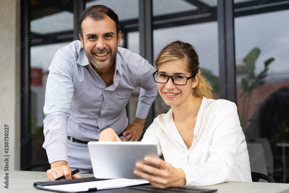 Portrait of happy business colleagues with digital tablet looking at camera and smiling. Young Caucasian woman wearing glasses sitting at table and mid adult man standing by. Team concept - obrazy, fototapety, plakaty 