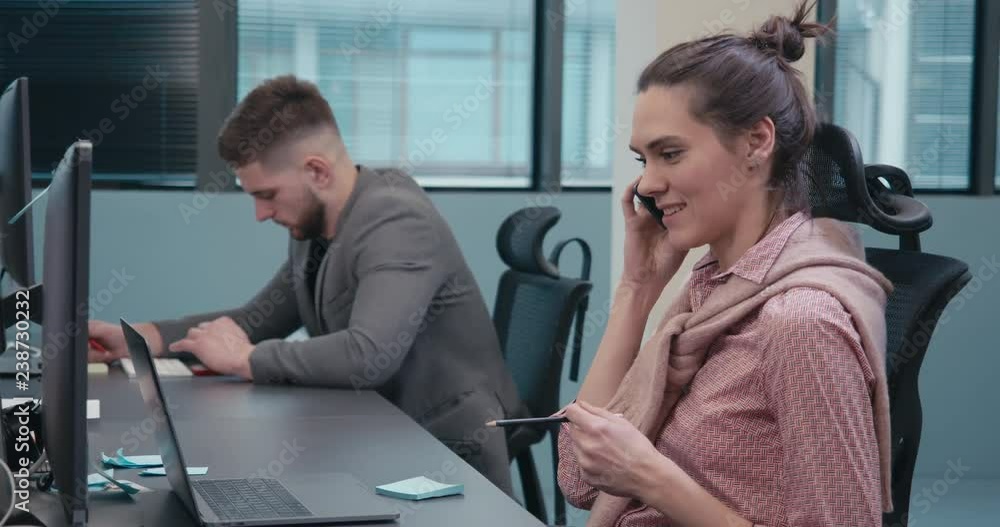 Happy young smiling Caucasian woman sitting at her desk in the office working and answering a phone call. 4K UHD Happy young smiling Caucasian woman sitting at her desk in the office working and answering a phone call. 4K UHD