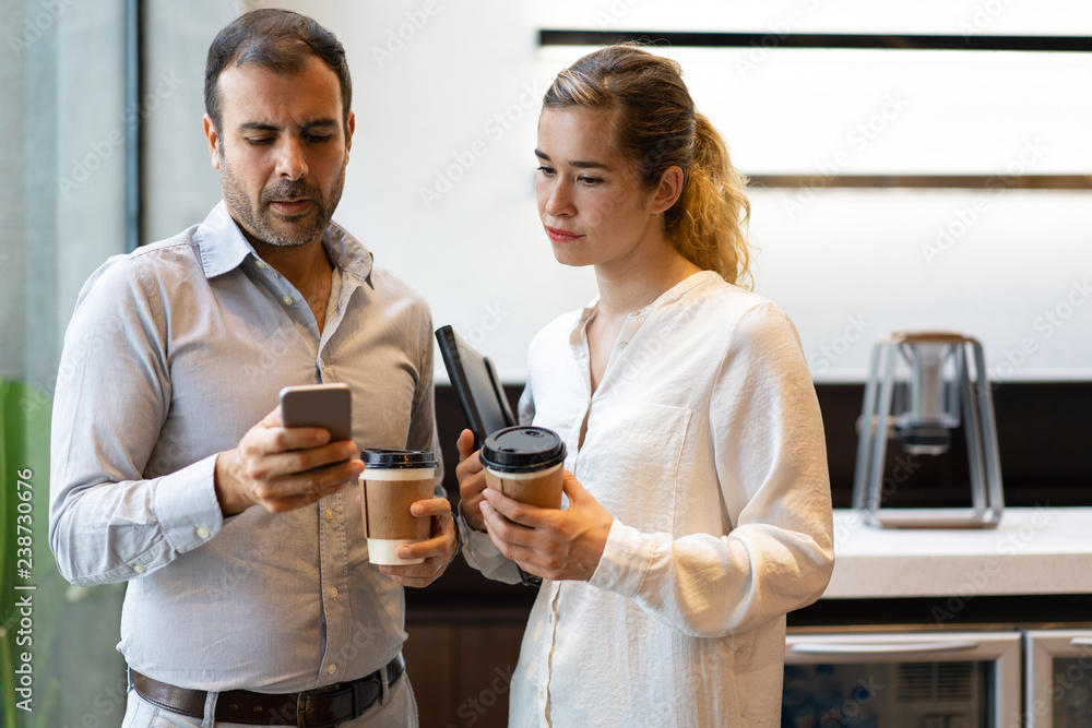 Serious businesspeople calling up colleague during coffee break. Young woman and mid adult bearded man standing with coffee cups at office kitchen and texting message on phone. Communication concept Serious businesspeople calling up colleague during coffee break. Young woman and mid adult bearded man standing with coffee cups at office kitchen and texting message on phone. Communication concept