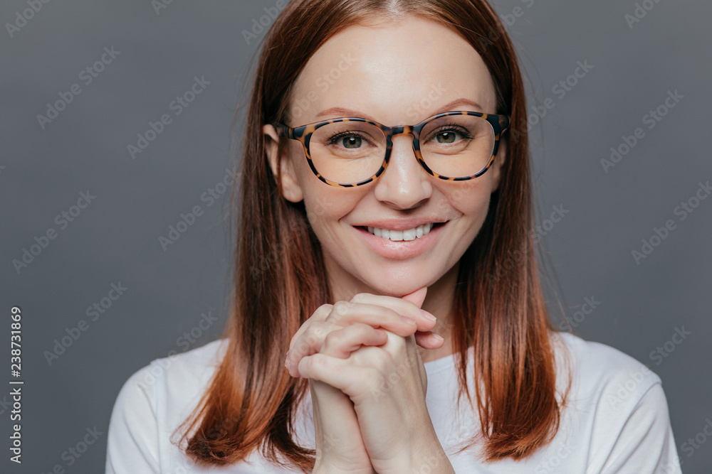 Close up shot of happy young European woman has pleasant smile, keeps hands together near chin, wears casual white clothes, isolated over grey background, being in high spirit, enjoys spare time