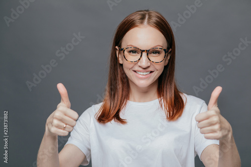 Glad positive woman with tender smile on face, has brown hair, raises two thumbs, demonstrates her approval, isolated over grey background, wears spectacles, wears casual white t shirt. Body language