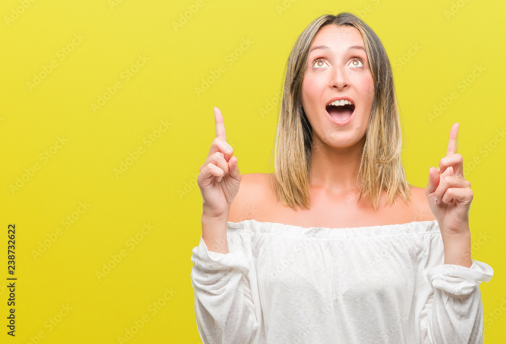 Young beautiful woman over isolated background amazed and surprised looking up and pointing with fingers and raised arms. Young beautiful woman over isolated background amazed and surprised looking up and pointing with fingers and raised arms.