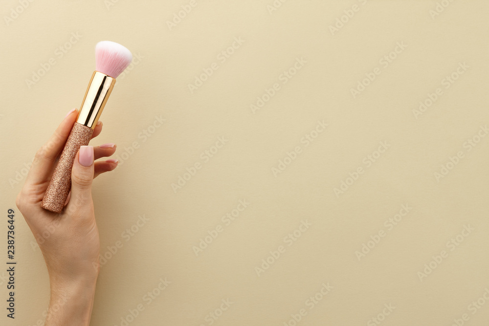 Woman's hand holding golden glossy make up brush with pink bristles on beige background, close up view, copy space Woman's hand holding golden glossy make up brush with pink bristles on beige background, close up view, copy space
