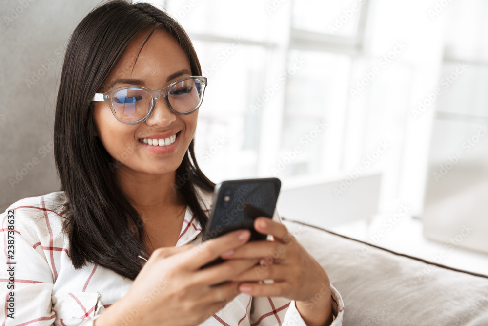 Photo of modern asian woman 20s holding smartphone, while lying at sofa in cozy apartment