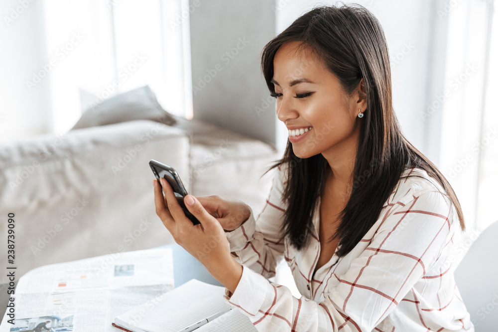 Image of brunette asian woman 20s using mobile phone, while sitting at table indoor and working with documents Image of brunette asian woman 20s using mobile phone, while sitting at table indoor and working with documents