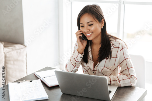 Image of joyous asian woman 20s talking on smartphone and working on laptop, while sitting at table indoor