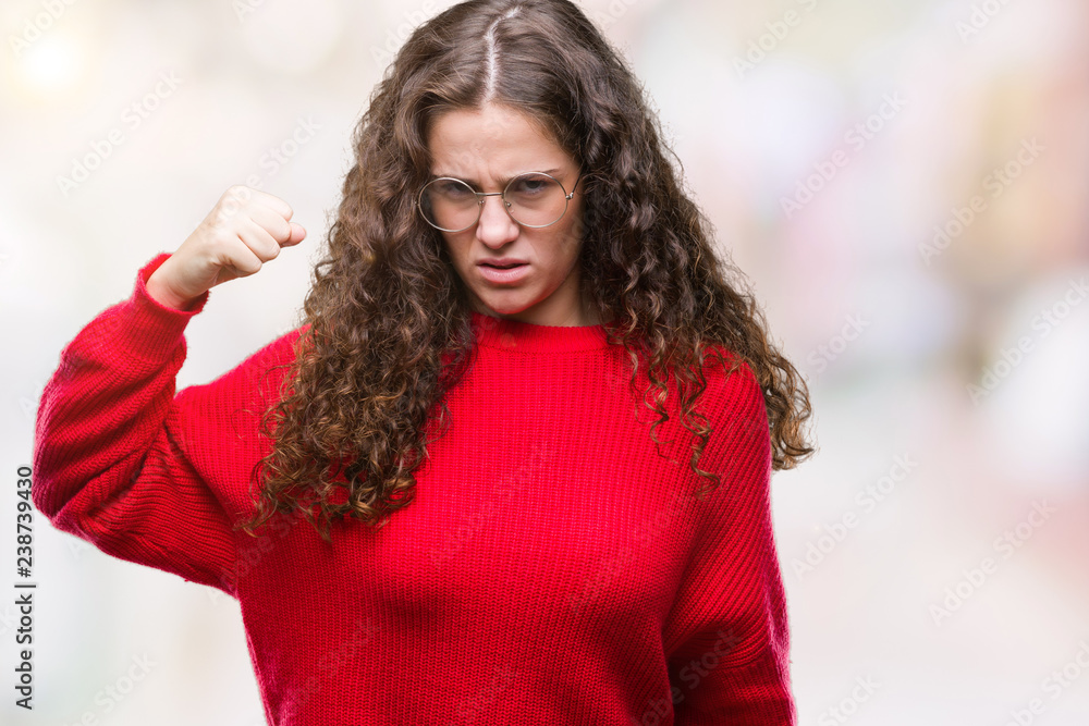 Beautiful brunette curly hair young girl wearing glasses and winter sweater over isolated background angry and mad raising fist frustrated and furious while shouting with anger. Rage and aggressive