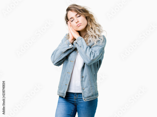 Beautiful young blonde woman wearing denim jacket over isolated background sleeping tired dreaming and posing with hands together while smiling with closed eyes.