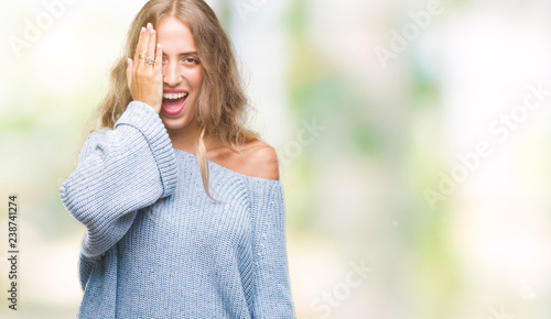 Beautiful young blonde woman wearing winter sweater over isolated background covering one eye with hand with confident smile on face and surprise emotion.