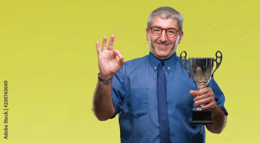 Handsome senior successful man holding trophy over isolated background doing ok sign with fingers, excellent symbol