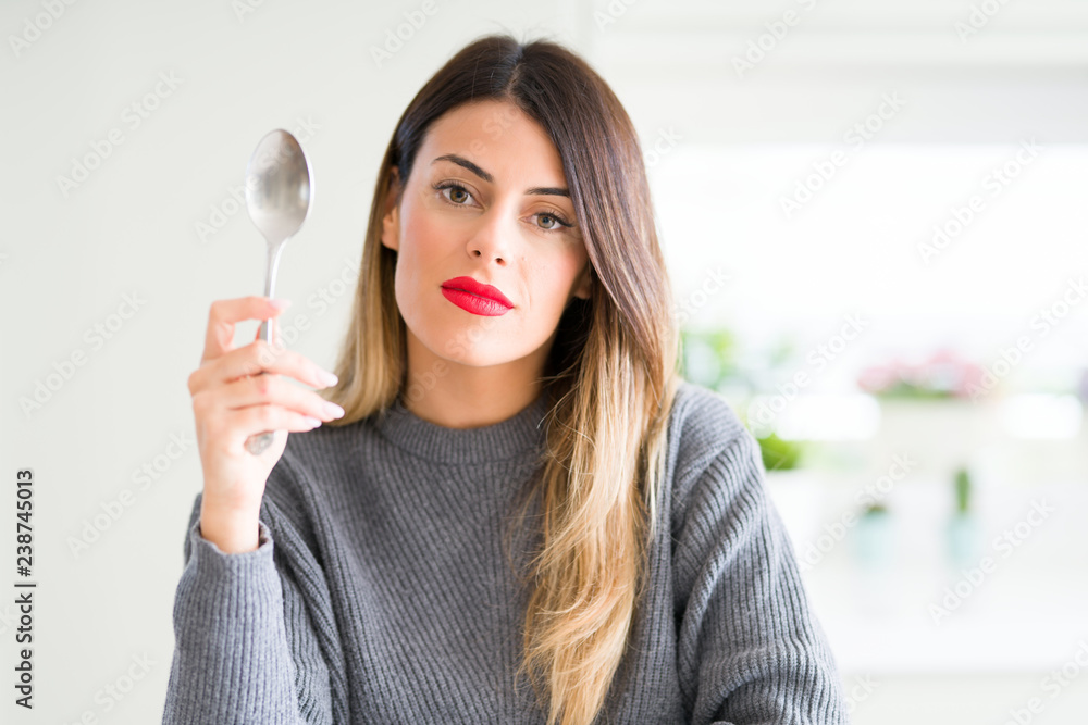 Young beautiful woman holding silver spoon at home with a confident expression on smart face thinking serious Young beautiful woman holding silver spoon at home with a confident expression on smart face thinking serious