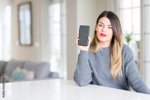 Young beautiful woman showing smartphone screen at home with a confident expression on smart face thinking serious