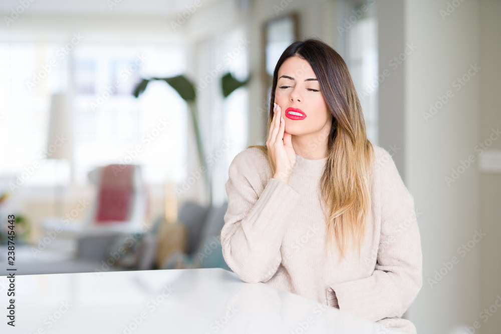 Young beautiful woman wearing winter sweater at home touching mouth with hand with painful expression because of toothache or dental illness on teeth. Dentist concept. Young beautiful woman wearing winter sweater at home touching mouth with hand with painful expression because of toothache or dental illness on teeth. Dentist concept.