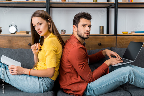 young couple sitting back to back on sofa with notebook and laptop