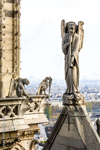 Stone statue of an angel with trumpet on the rooftop of Notre-Dame de Paris cathedral and three chimeras on the towers gallery overlooking the buildings of the city vanishing in the mist.
