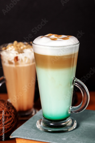 Coffee latte macchiato with mint syrup and whipped cream, in high transparent glasses, on a wooden table, on a dark background with cakes