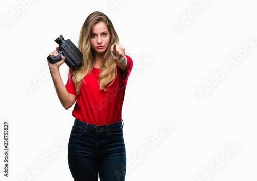 Young beautiful blonde woman filming using vintage camera over isolated background pointing with finger to the camera and to you, hand sign, positive and confident gesture from the front