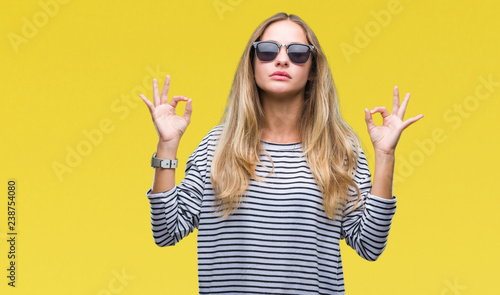 Young beautiful blonde woman wearing sunglasses over isolated background relax and smiling with eyes closed doing meditation gesture with fingers. Yoga concept.