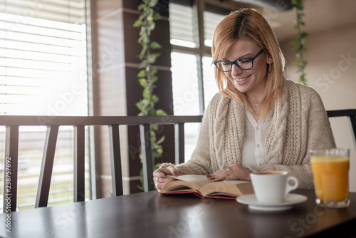 Smiling girl with glasses reading book, drinking juice and coffee in cafe.