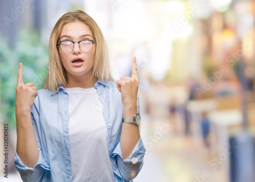 Young caucasian business woman wearing glasses over isolated background amazed and surprised looking up and pointing with fingers and raised arms.