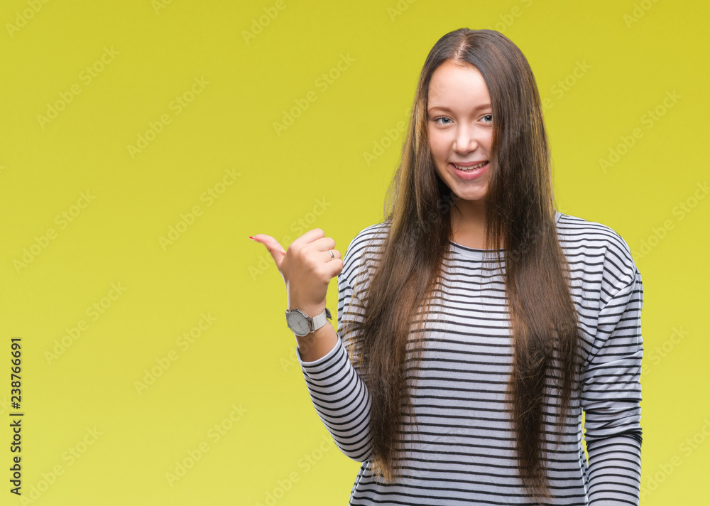 Young beautiful caucasian woman over isolated background smiling with happy face looking and pointing to the side with thumb up. Young beautiful caucasian woman over isolated background smiling with happy face looking and pointing to the side with thumb up.