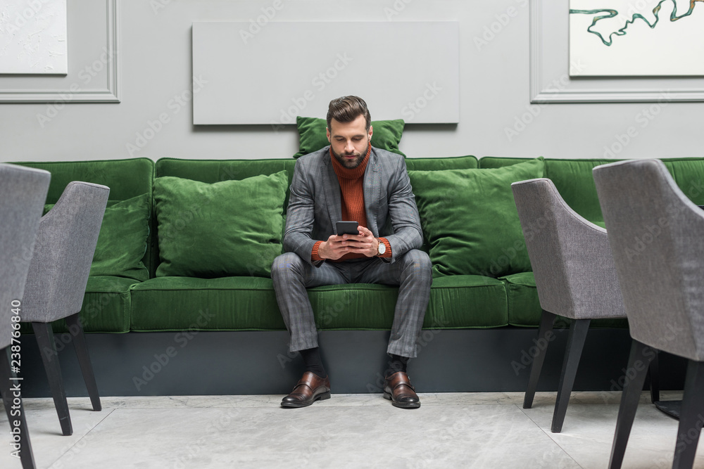 serious handsome man in formal wear sitting on green sofa and using smartphone