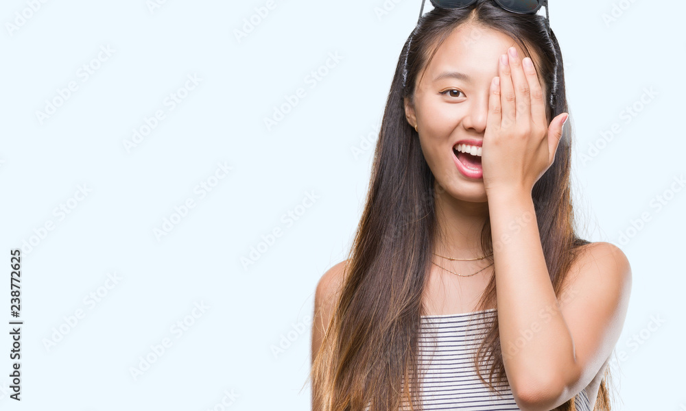 Young asian woman wearing sunglasses over isolated background covering one eye with hand with confident smile on face and surprise emotion. Young asian woman wearing sunglasses over isolated background covering one eye with hand with confident smile on face and surprise emotion.